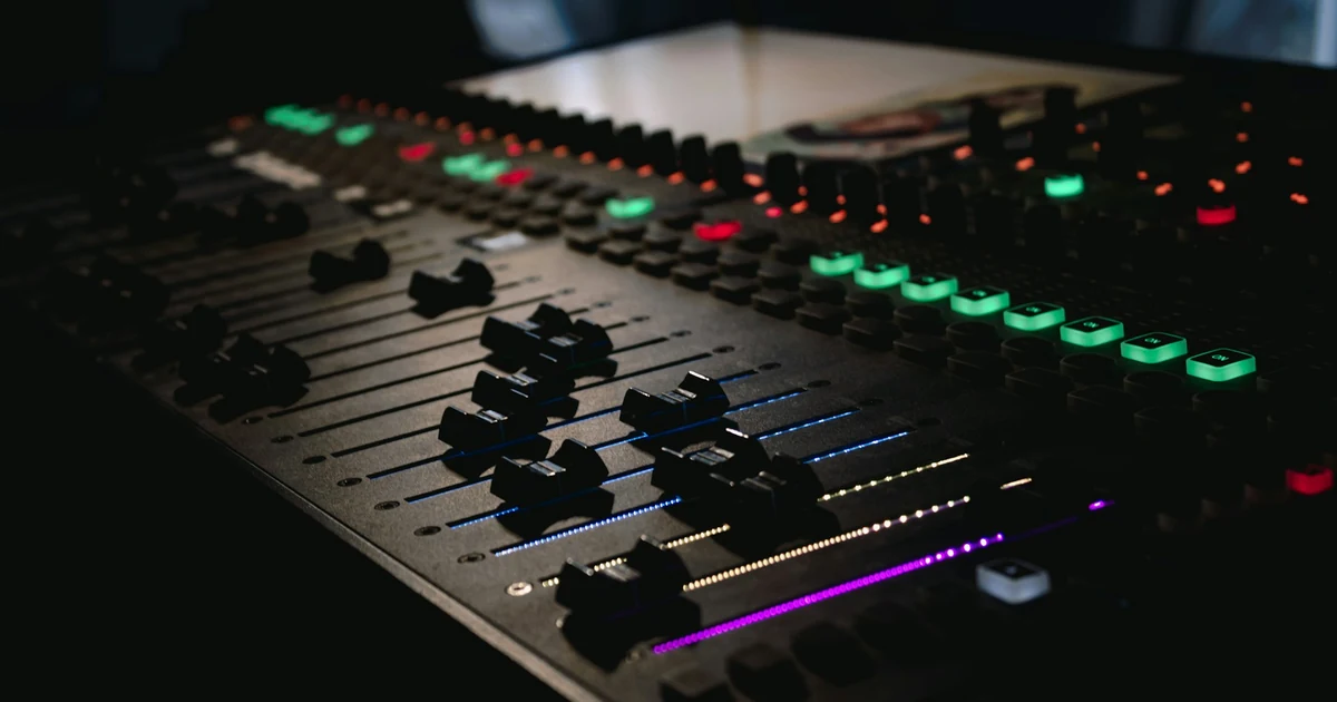 A dark audio mixing console with rows of faders set to different levels and glowing channel lights